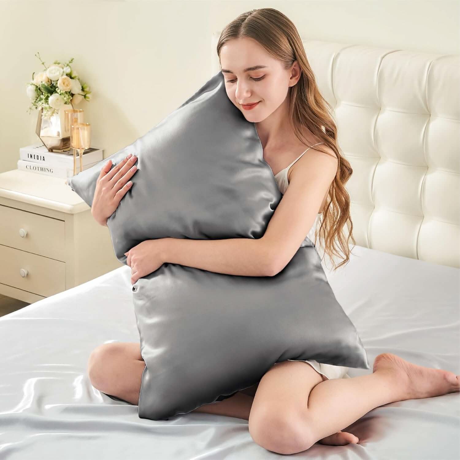 Smiling woman sitting on a bed hugging a silver silk pillowcase, emphasizing its soft and luxurious feel