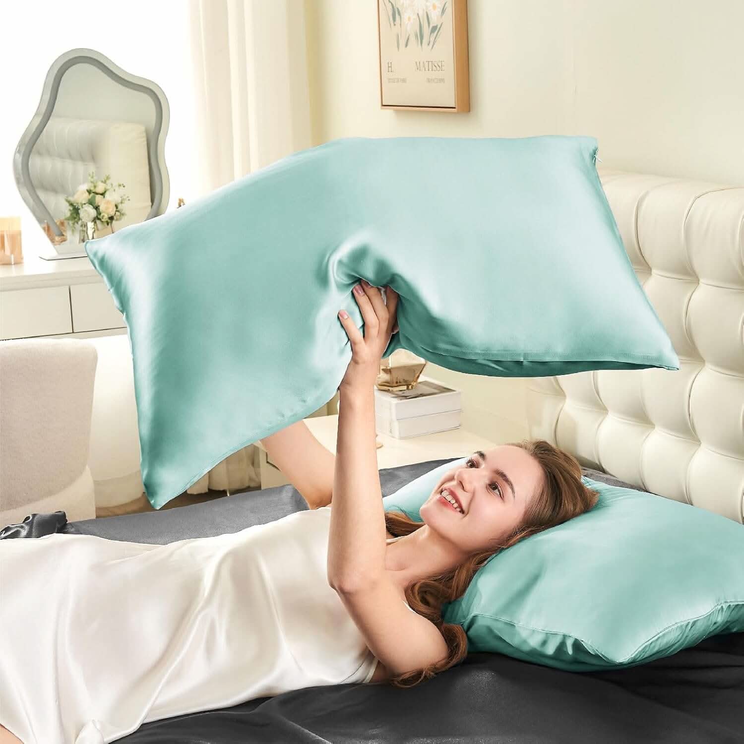 Woman lying on a bed joyfully tossing a light blue silk pillowcase in the air, highlighting its soft and smooth texture