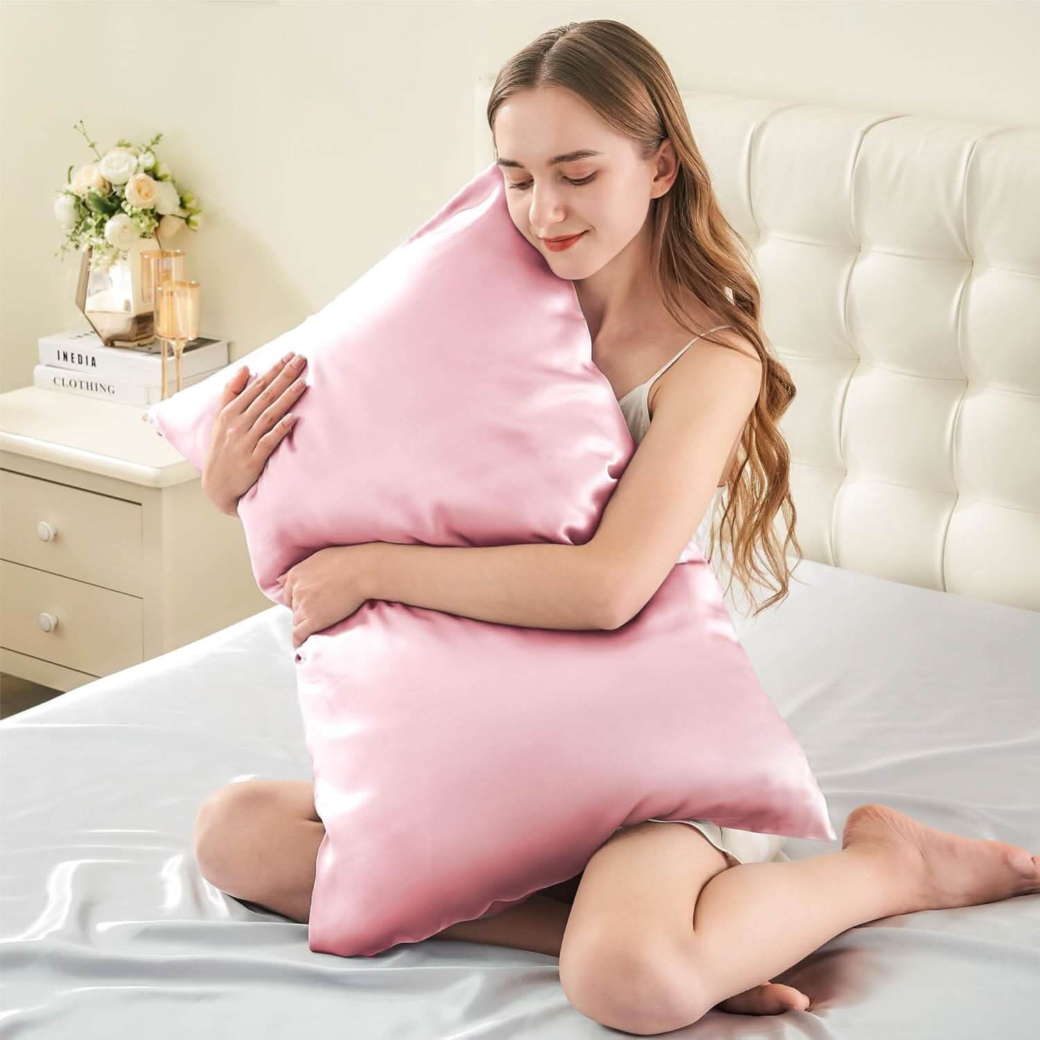 Smiling woman sitting on a bed hugging a pink silk pillowcase, emphasizing its soft and luxurious feel