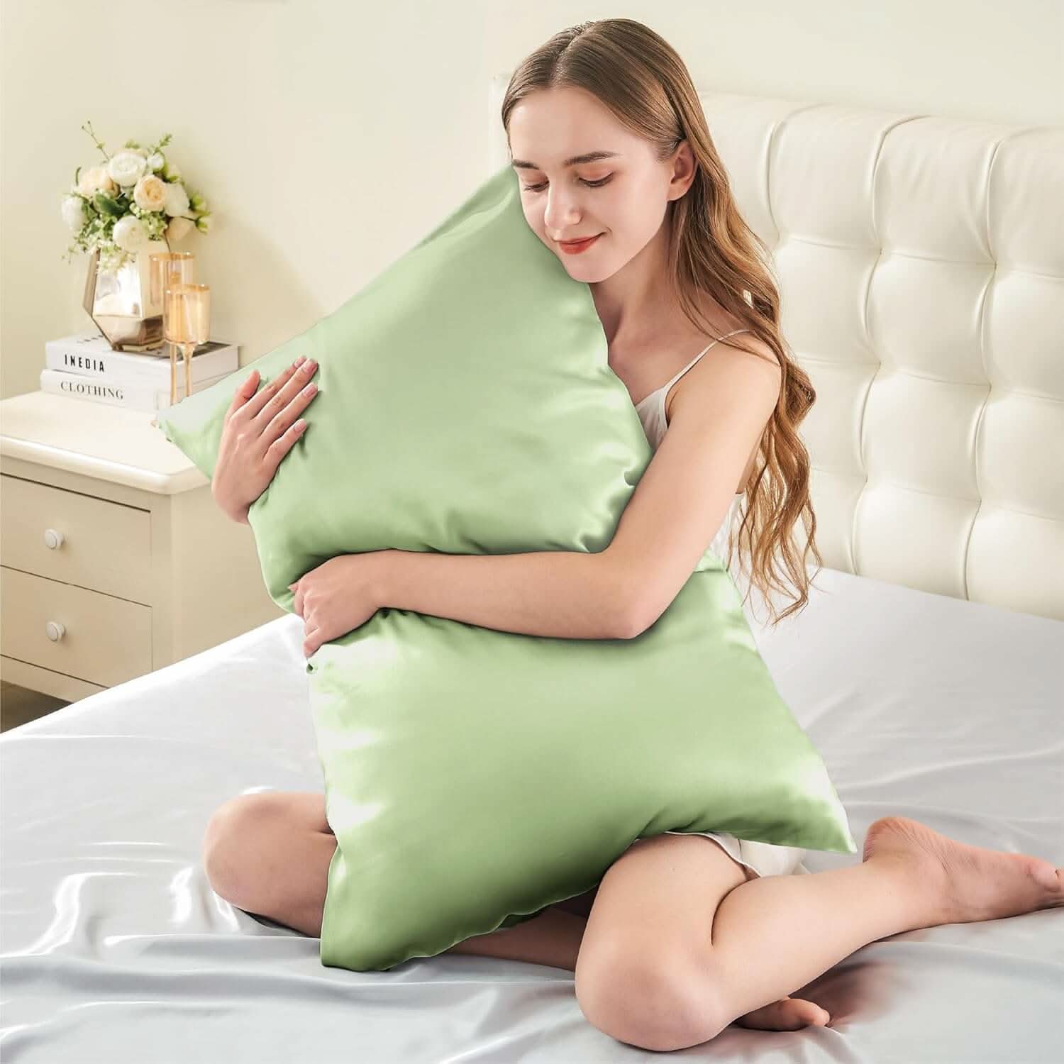 Smiling woman sitting on a bed hugging a sage green silk pillowcase, emphasizing its soft and luxurious feel