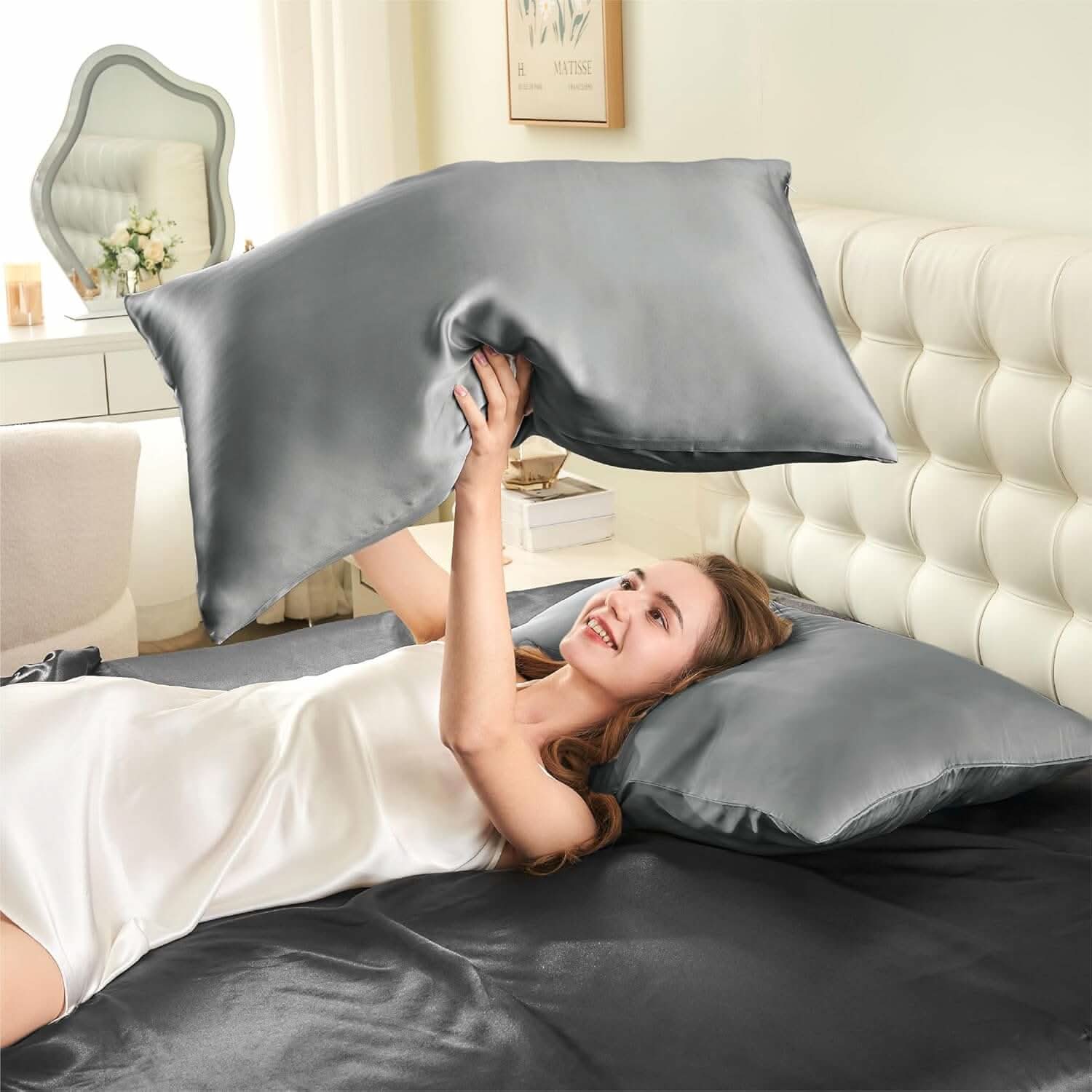 Woman lying on a bed joyfully tossing a silver silk pillowcase in the air, showcasing its soft and smooth texture.