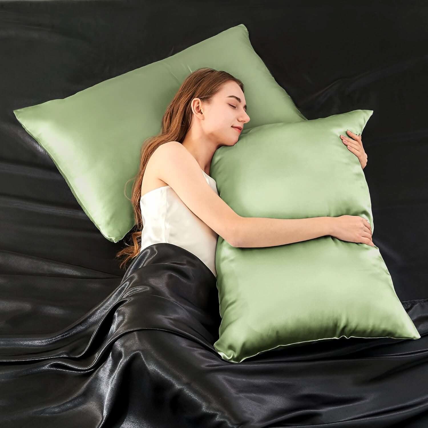 Woman lying on a bed hugging a sage green silk pillowcase, highlighting its smooth and luxurious texture