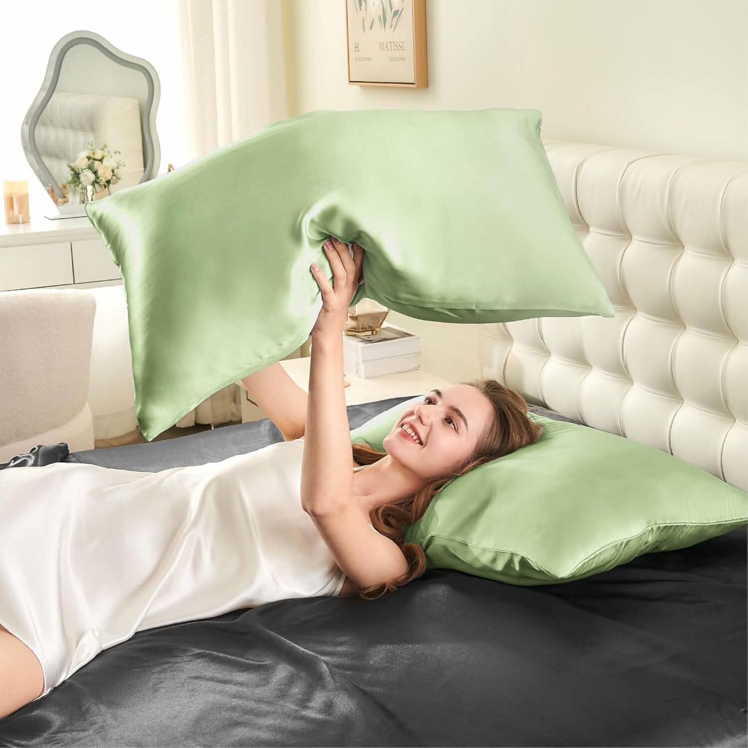 Woman lying on a bed joyfully tossing a sage green silk pillowcase in the air, showcasing its soft and smooth texture.