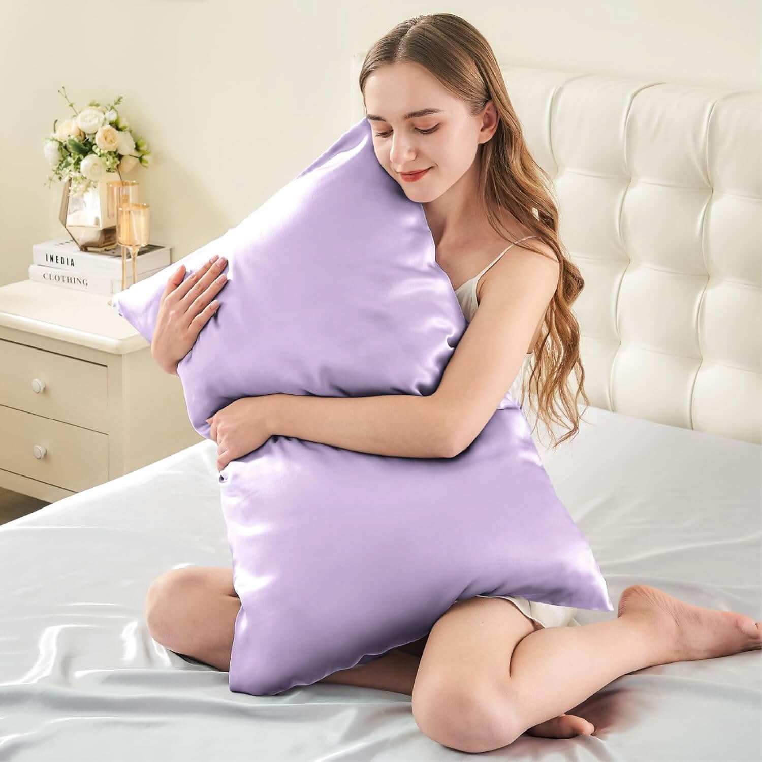 Smiling woman sitting on a bed hugging a lavender silk pillowcase, emphasizing its soft and luxurious feel.
