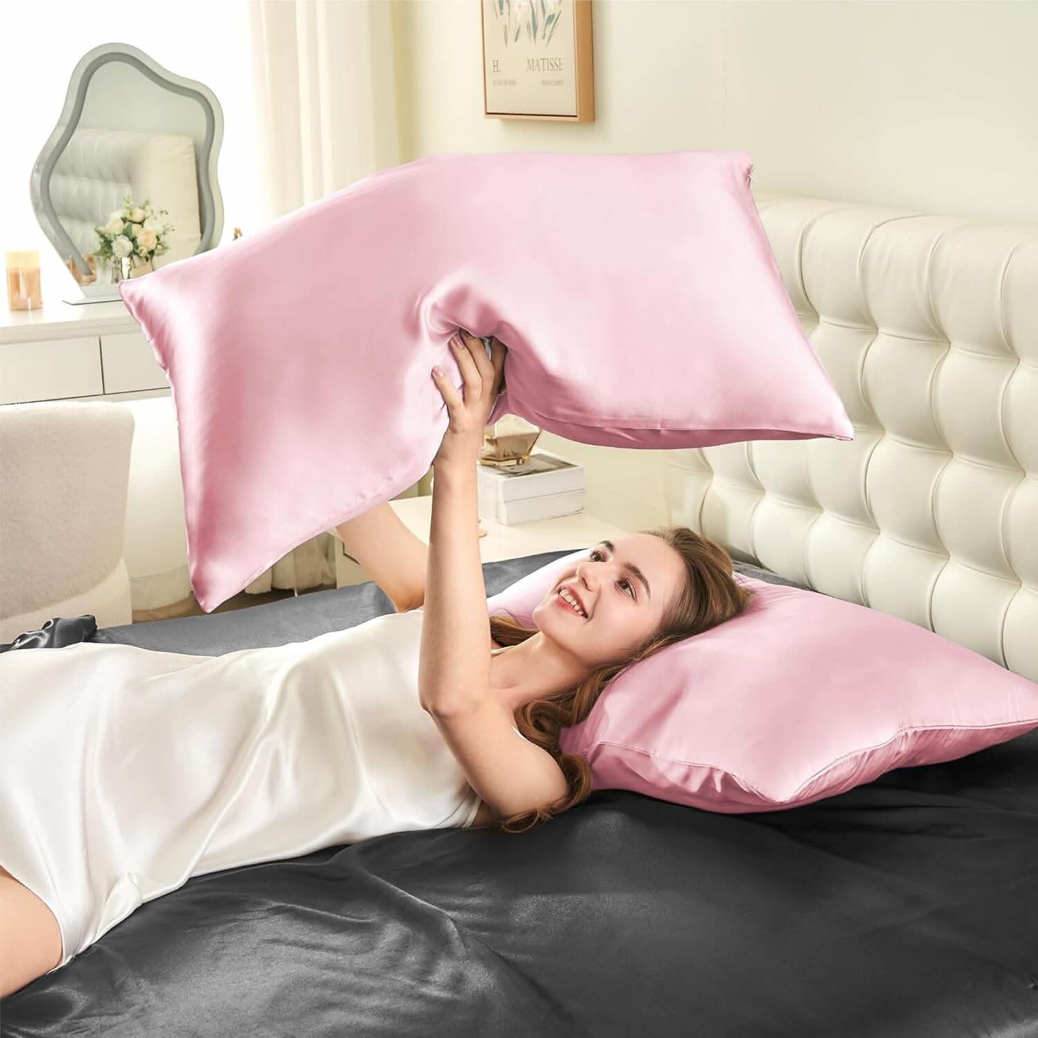 Woman lying on a bed joyfully tossing a pink silk pillowcase in the air, showcasing its soft and smooth texture