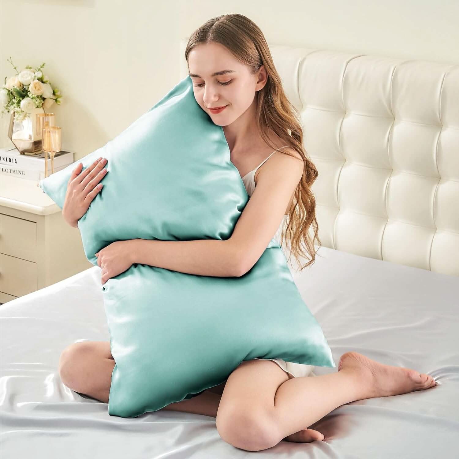 Smiling woman sitting on a bed hugging a light blue silk pillowcase, emphasizing its soft and luxurious feel.