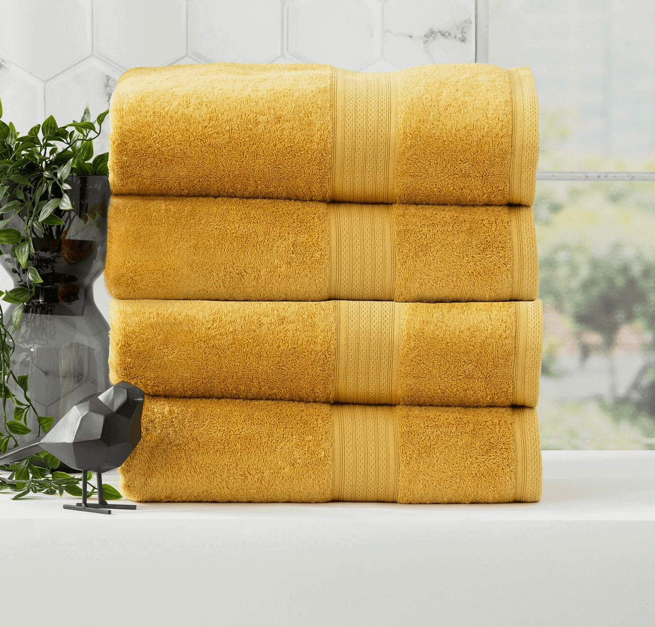 Stack of four yellow bath towels next to a geometric bird sculpture and a green plant in a bathroom.