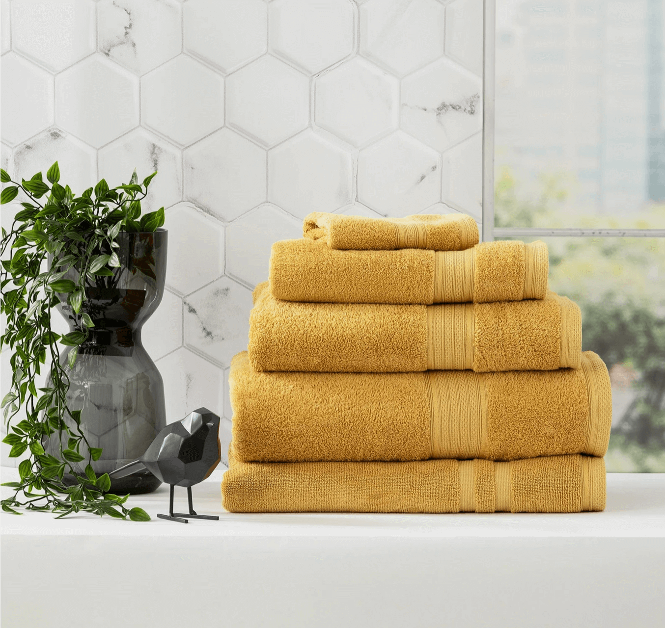 A stack of folded yellow towels next to a black vase with green plant and decorative bird on a white countertop